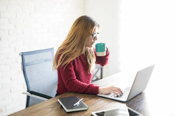 年轻的白种女人喝咖啡和使用笔记本电脑在家里办公室 — 图库照片