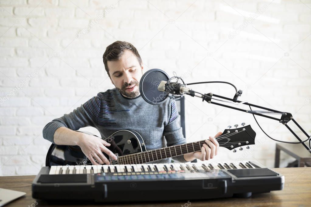 Man in his 30s playing live music on online radio, singing song into mic and playing guitar.