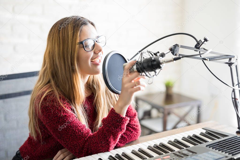 Cute young female sitting with a piano and singing into the microphone, podcaster on online studio.