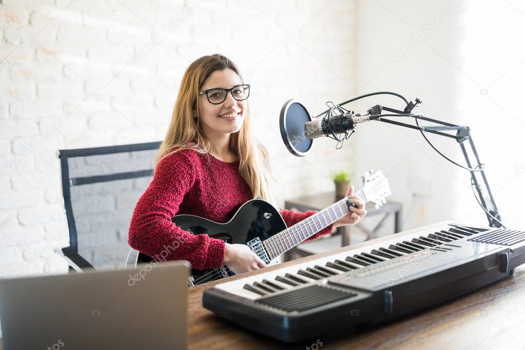 Portrait of cute young caucasian female musician sitting at table with electric piano and playing guitar