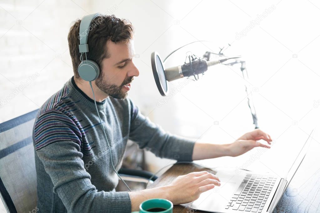 Profile view of handsome young latin man using laptop and taking into microphone, hosting online radio.