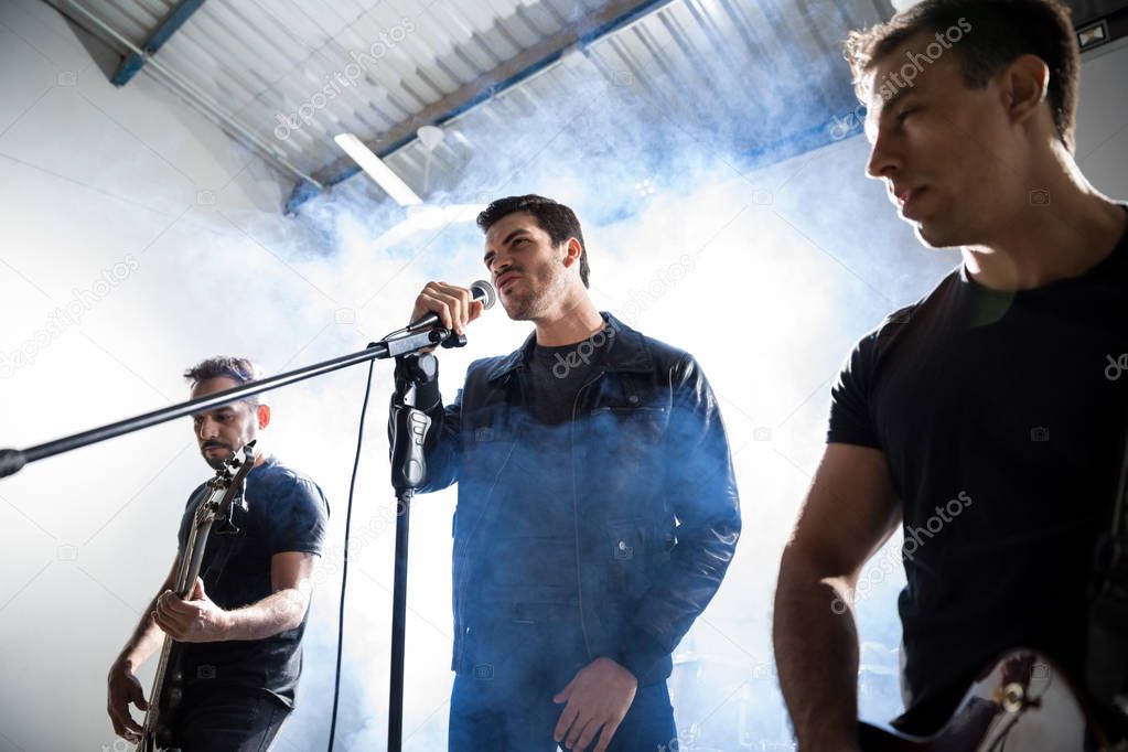 Male lead singer with two guitar players performing music on stage with smoke and light at back