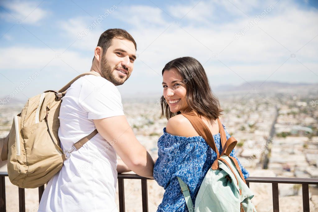 Cheerful young couple at vacation wearing casuals and backpack looking at camera with cityscape background