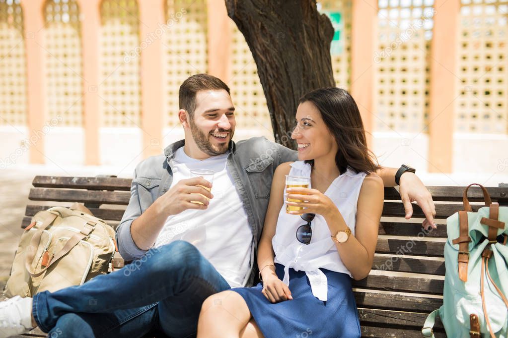 Beautiful couple looking at each other while holding wine glasses sitting on bench with bags besides them