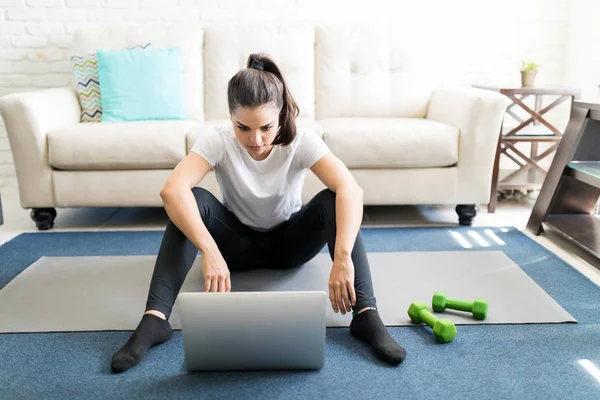 年轻的拉丁妇女坐在运动垫在家和观看健身教程在线上的笔记本电脑 — 图库照片