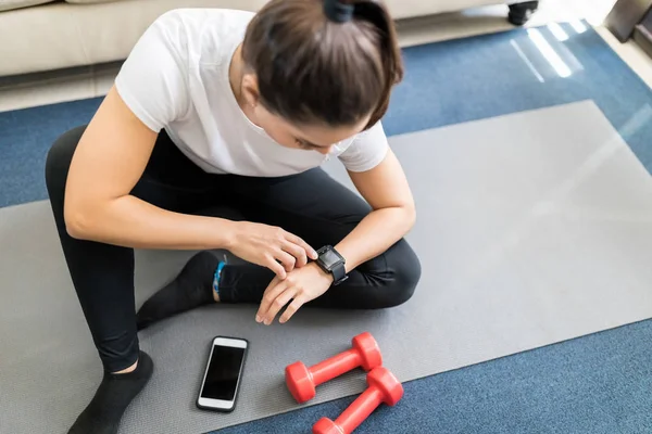 年轻女子坐在瑜伽垫在客厅和设置 Smartwatch 前锻炼 — 图库照片