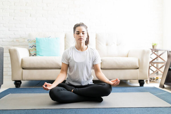 young hispanic woman meditating in lotus yoga pose in living room