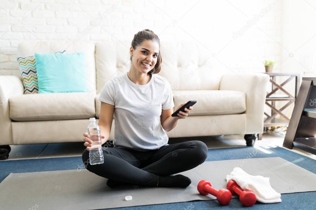 young hispanic woman sitting on exercise mat with smartphone and water bottle