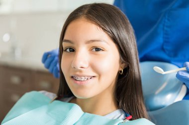 Dişçi klinikte ayakta iken sandalye üzerinde oturan diş telleri ile gülümseyen genç kız portresi 