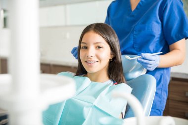 Dişçi klinikte ayakta iken sandalye üzerinde oturan diş telleri ile gülümseyen genç kız portresi 