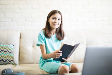 Dizüstü bilgisayar ile oturma odasında kanepede kitap ve kulaklık masada otururken ev ödevi Tamamlanıyor gülümseyen liseli kız
