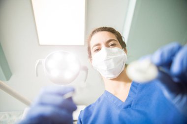 Işıklı diş kliniği aracıyla çalışırken Ameliyat maskesi giyen kadın diş hekimi düşük açılı portresi 