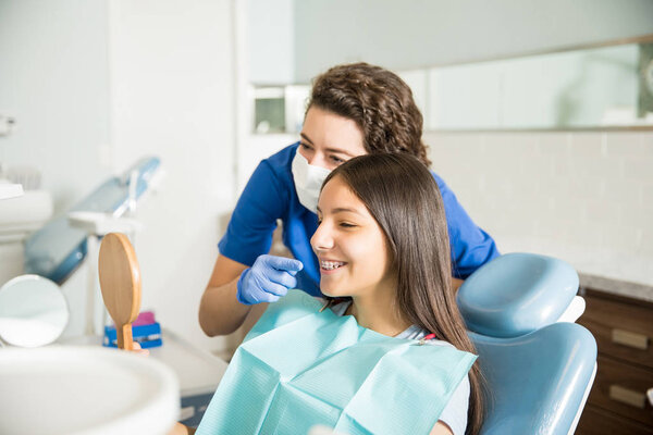 Female dentist showing braces to teenage girl in mirror at dental clinic