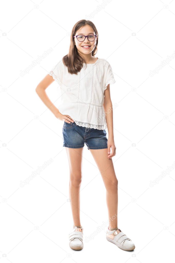Full length portrait of cute little girl in casual clothes and wearing spectacles looking at camera and smiling while standing isolated on white background