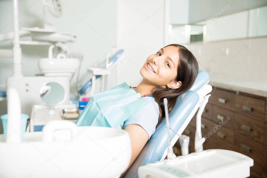 Portrait of smiling teenage girl with braces sitting on chair at dental clinic