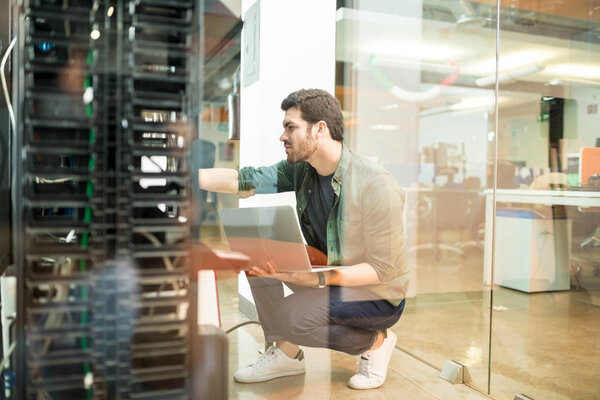 Portrait of happy young male network engineer with laptop in hand working in datacenter 