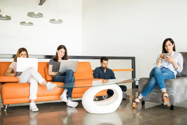 四位年轻的拉丁人坐在办公室休息室使用笔记本电脑和移动电话 同时放松在休息期间 — 图库照片