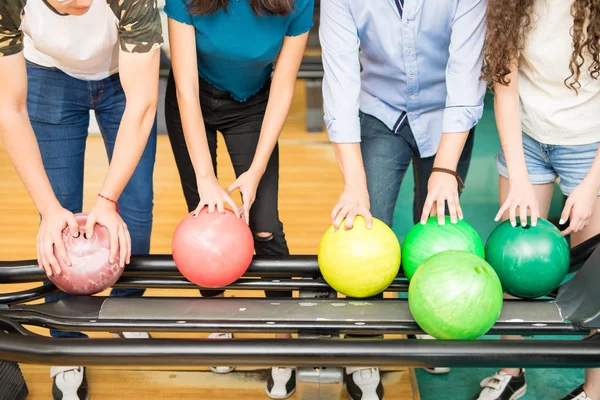 Teenage friends with multicolored balls at rack in bowling alley at club