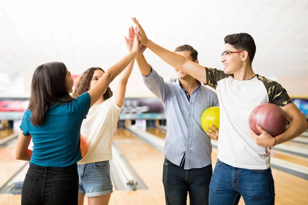 Male and female teenage friends giving high-five while holding bowling balls at alley in club