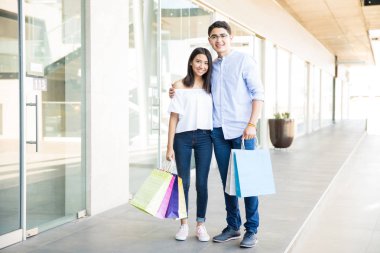 Gülümseyen genç sevgili ile renkli alışveriş ayakta ve alışveriş merkezi koridorda kameraya bakarak torbaları 