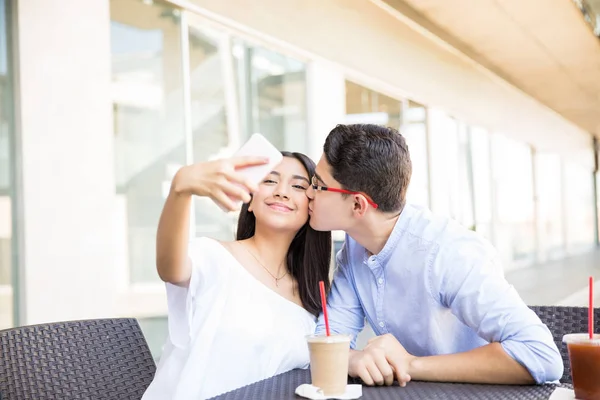 十几岁的女孩采取自拍 而男朋友亲吻她的脸颊在购物中心咖啡馆 — 图库照片