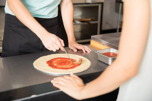 Closeup of chef hands applying tomato puree ketchup on freshly baked pizza bread in front of customer at counter