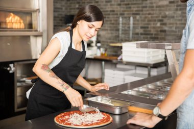 Paslanmaz Kaşık peynir pizza üzerinde temel yağmurlama tutarak siyah önlük giyen genç kadın ile domates sosu katmanlı