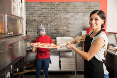 Şef giyen üniforma kamera ve pizza dükkanı arka planda çalışan personel bakarken pişirme için modern fırında ham pizza koyarak gülümseyen