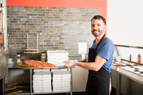 Portrait of smiling chef wearing black apron uniform holding big iron pizza peel with freshly baked pizza removed from oven in pizza shop kitchen counter