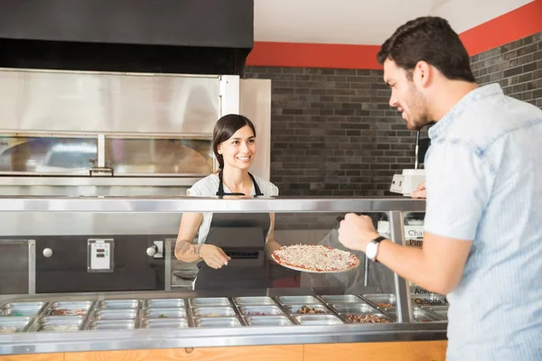 在比萨店的顾客选择意大利比萨在比萨饼商店与女厨师身穿黑色围裙和举行比萨饼顶部番茄酱和干酪奶酪后厨房柜台 — 图库照片