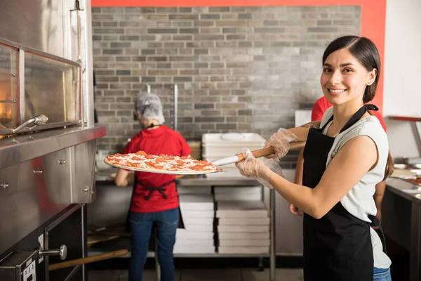 微笑的厨师穿着制服把生比萨饼放在现代烤箱里烘烤 同时看着相机和在披萨店后台工作的工作人员 — 图库照片