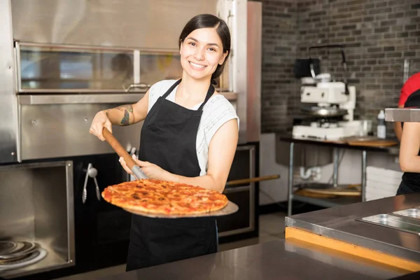 在比萨店工作的开朗的年轻妇女提出新鲜烘焙比萨的皮站在柜台后面 — 图库照片
