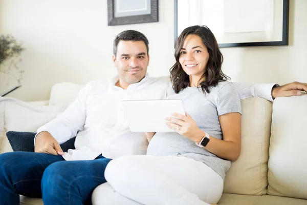 带数字平板电脑的自信怀孕夫妇的肖像坐在沙发上在家 — 图库照片