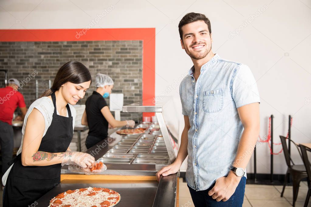 Cheerful man standing at kitchen counter at pizza shop with beautiful woman in black apron and wearing disposable gloves adding pepperoni slices to cheese grated pizza