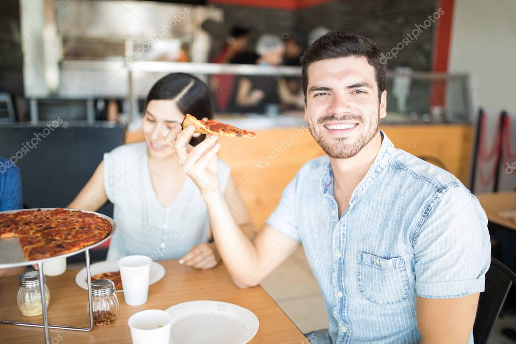 Attractive young man sitting in indoor pizza shop with friends enjoying pizza party while holding pizza slice and looking at camera