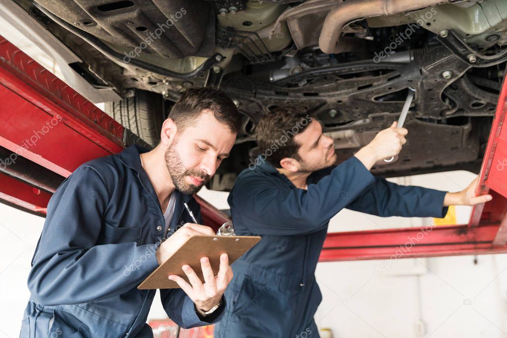 Mid adult mechanic taking notes while coworker repairing car in garage
