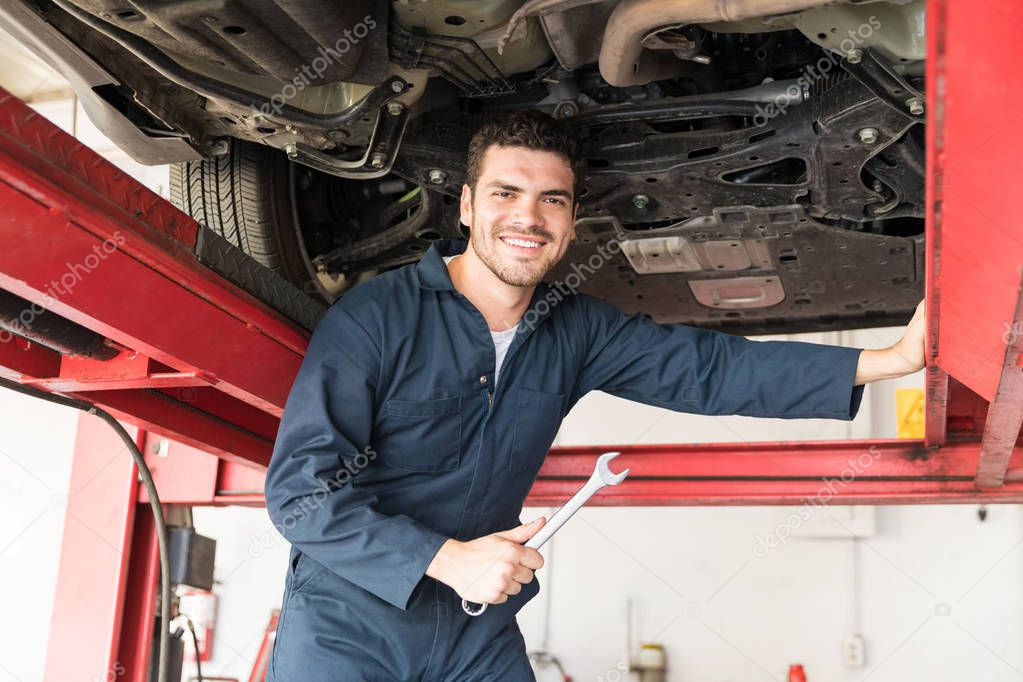 Portrait of smiling maintenance engineer holding wrench while standing under car in repair shop