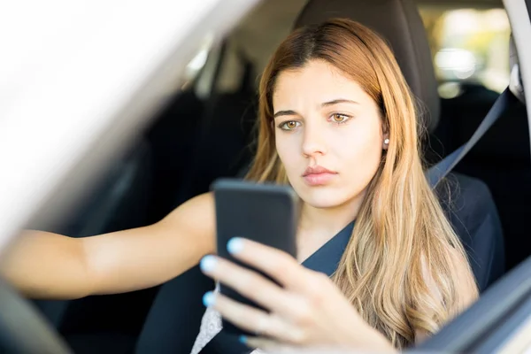 テキスト メッセージを読むための魅力的な若い女性彼女の車の運転席に座って 携帯電話を見ての肖像画 — ストック写真