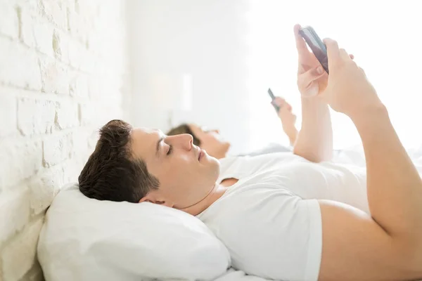 成人中年男子的侧面查看智能手机短信 而躺在床上的女人在家 — 图库照片