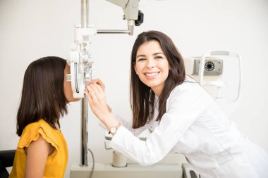 Portrait of happy beautiful woman doing eye test of girl child in optical store while looking at camera clipart