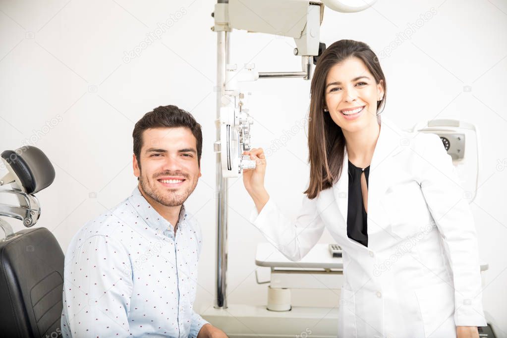 Cheerful man sitting on chair with beautiful optician standing while dong eye test using phoropter in optical laboratory