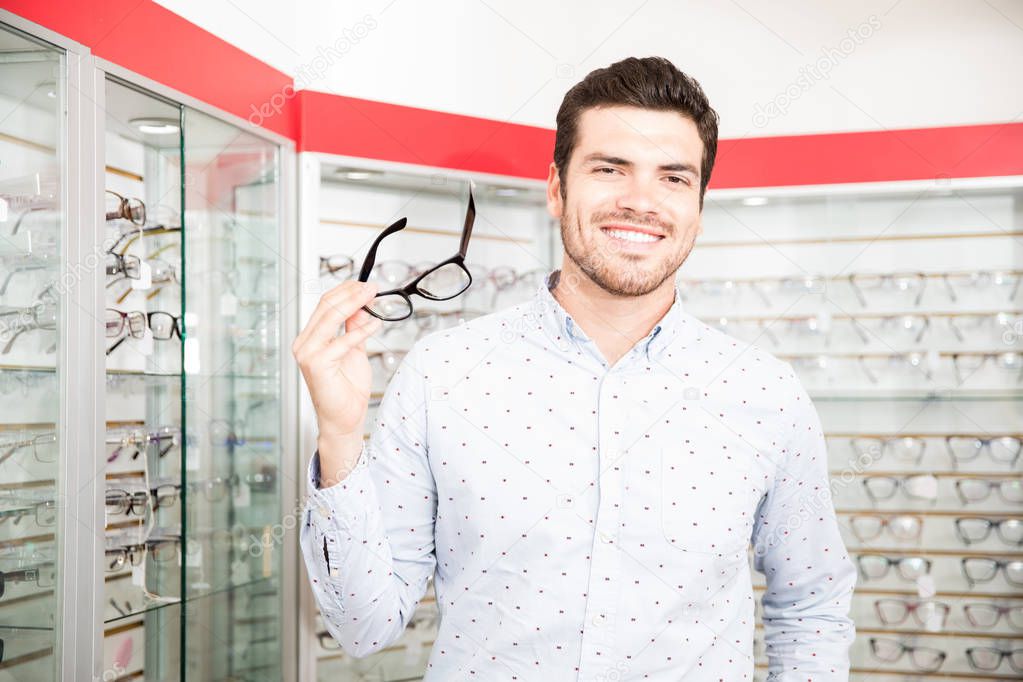 Handsome smiling man trying black spectacles with retro look standing in front of display shelves in optics sop while looking at camera