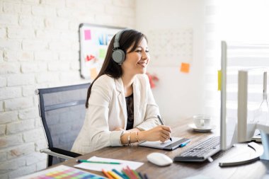 Bazı tasarımlar hazırlamak için grafik tablet ve bilgisayar kullanarak kulaklık giyen mutlu genç latin kadın portresi