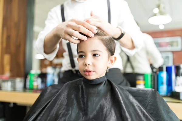 Cute boy looking away while young hairdresser styling his hair in shop