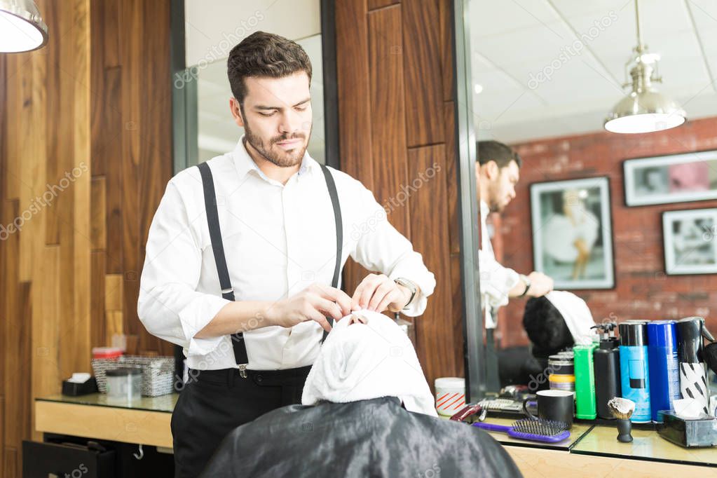 Barber applying hot towel on client's face before giving him a shave in shop