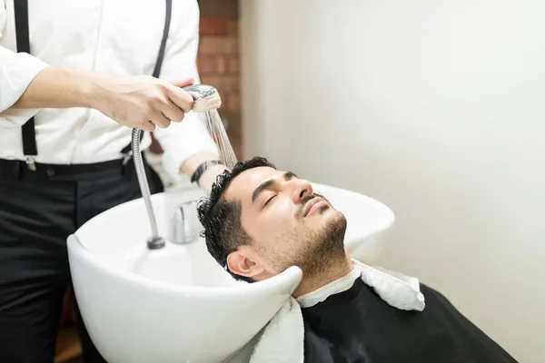 放松的年轻人 眼睛闭上倚在水槽 而理发师洗他的头发在沙龙 — 图库照片