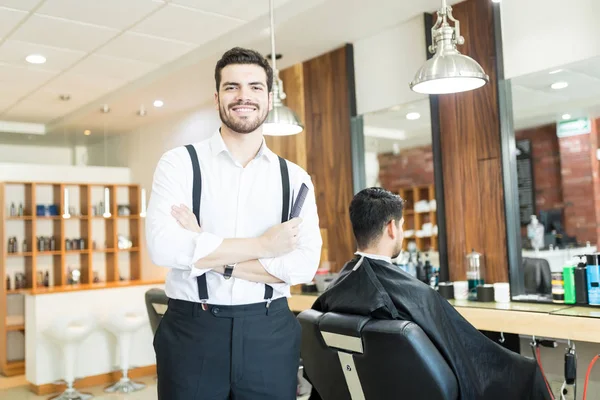 凉爽的理发师与手臂交叉站在椅子上的客户坐在沙龙 — 图库照片
