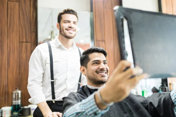 Attractive man holding mirror while looking at his new haircut from barber in salon