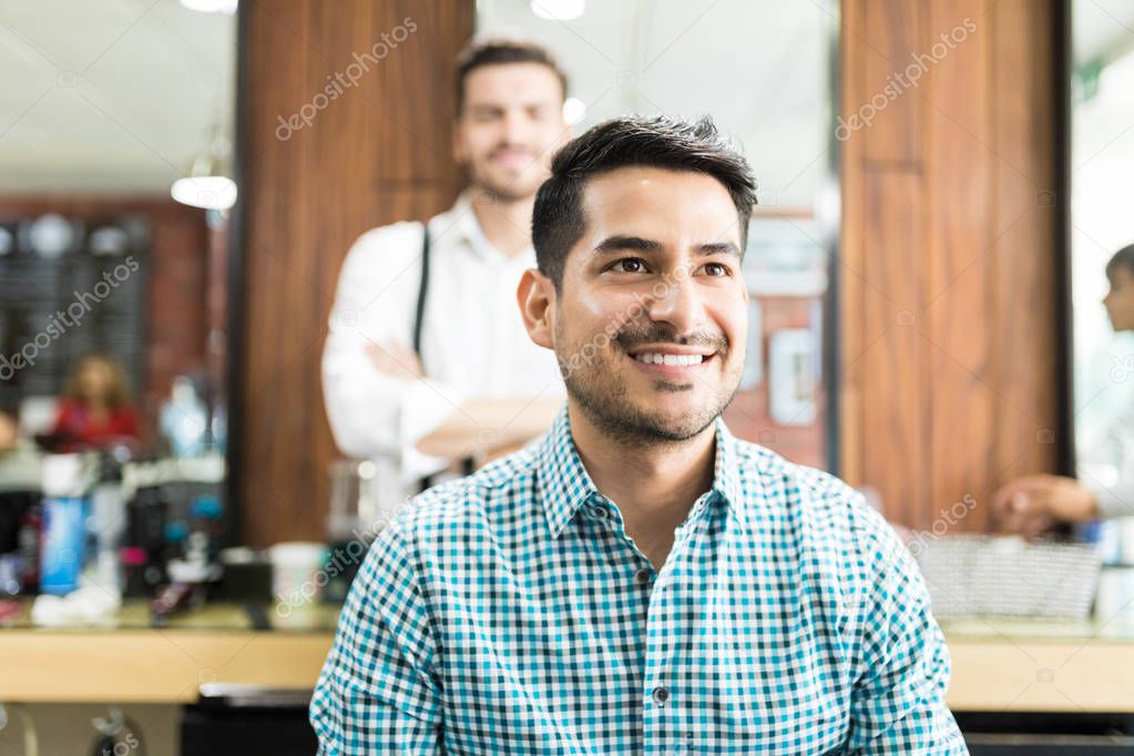 Smiling young man looking away after haircut with barber in background at salon