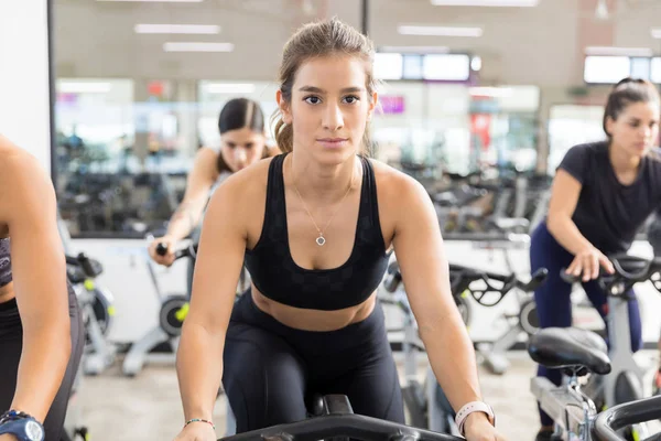 在健身俱乐部使用固定式自行车的坚定的年轻妇女在运动衫 — 图库照片
